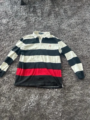 Randig rugbytröja från Polo Ralph Lauren - Klassisk långärmad rugbytröja från Polo Ralph Lauren med breda svarta, vita och en röd rand. Tröjan har krage, knäppning vid halsen och ikonisk liten logga på bröstet. Tillverkad i mjuk bomull för en avslappnad men snygg look.
