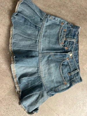 Kort jeanskjol med volangkant - Säljer en ljusblå jeanskjol med volangkant och klassiska fickor fram. Kjolen har en kort modell och råa kanter nertill för en trendig look. Perfekt för dig som gillar denim och vill ha en cool, avslappnad stil. 