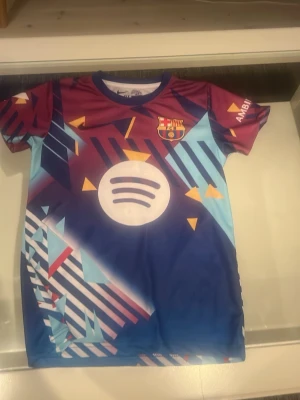 Barcelona matchtröja Lamine Yamal - Snygg Barcelona matchtröja med Lamine Yamal och nummer 10 på ryggen. Tröjan har ett färgstarkt geometriskt mönster i blått, vinrött, vitt och gult samt tryckta loggor från Spotify och UNHCR. Korta ärmar och rund hals, perfekt för fotboll eller träning.