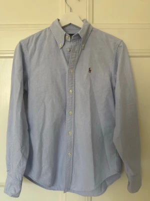 Ljusblå skjorta från Ralph Lauren - Klassisk ljusblå skjorta från Ralph Lauren med button-down krage och broderad logga på bröstet. Skjortan har långa ärmar, vita knappar och är tillverkad i mjuk bomull. Perfekt till jeans eller chinos för en clean look.