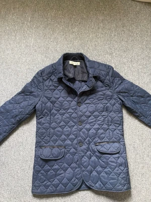 Marinblå quiltad jacka Close by Denim - Snygg marinblå quiltad jacka från Close by Denim. Jackan har knäppning med tre knappar, två framfickor med lock och en klassisk krage. Materialet är lätt vadderat och ger en stilren look. Perfekt för dig som gillar en modern och avslappnad stil.