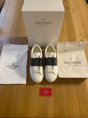 Valentino Opens - Snygga vita sneakers från Valentino Garavani opens med ett brett svart band över snörningen. Skorna har klassisk låg profil, vita skosnören och diskret logga på plösen. Tillverkade i slätt läder för en clean och exklusiv look.