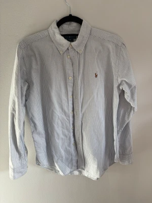 Randig skjorta från Polo Ralph Lauren - Klassisk vit och blå randig skjorta från Polo Ralph Lauren med broderad logga på bröstet. Skjortan har knappar framtill, button-down krage och långa ärmar. Tillverkad i mjuk bomull för en bekväm känsla.