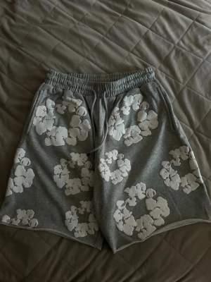 Grå shorts med vita blommor - Säljer ett par grå shorts med vita blommor i ett stort, tydligt mönster. Shortsen har resårmidja med snörning och är tillverkade i mjukt material, perfekta för avslappnade dagar. Bakficka och rymlig passform för extra komfort.