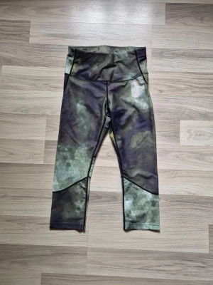 Camo träningsleggings med hög midja - Snygga träningsleggings med camouflage-mönster i gröna och mörka toner. De har hög midja och är tillverkade i ett stretchigt syntetmaterial, perfekta för träning eller chill. 7/8-längd och coola detaljer på benen.