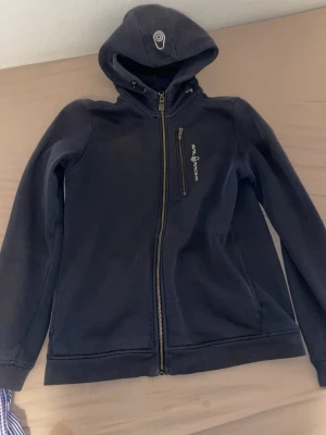 Marinblå hoodie från Sail Racing - Snygg marinblå hoodie från Sail Racing med dragkedja framtill och en bröstficka med logga. Huva med dragsko och broderad detalj. Tillverkad i mjukt material, perfekt för chill dagar eller när du vill ha en sportig look. Finns ett par olje fläck 