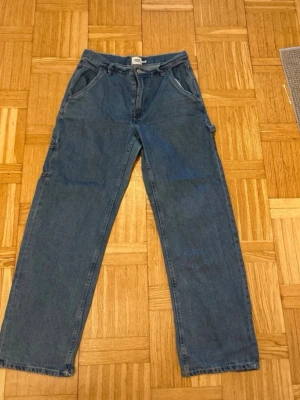 Blå raka jeans med verktygsficka - Ett par blå jeans med rak passform och verktygsficka på sidan. Klassisk femficksmodell med dragkedja och knapp i midjan. Jeansen har en avslappnad look och är tillverkade i denim. Perfekta för en casual stil.