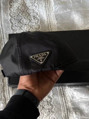 Prada keps  - Snygg svart keps från Prada i glansigt nylonmaterial med det ikoniska trekantiga metalloggan på sidan. Klassisk böjd skärm och justerbar rem bak. Perfekt accessoar för en trendig och stilren look.