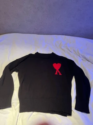 Ami Paris tröja - Snygg svart stickad tröja med ett stort rött hjärta och bokstaven A på bröstet. Tröjan har rund halsringning, ribbade muddar och är gjord i ett tjockare stickat material. Perfekt för dig som gillar stilrena plagg med en twist.