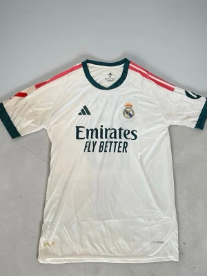 Real Madrid matchtröja vit - Säljer en vit Real Madrid matchtröja från Adidas med mörkgröna detaljer och rosa ränder på axlarna. Tröjan har klubbmärke, Adidas-logga och Emirates Fly Better-tryck på bröstet. Tillverkad i lätt och ventilerande material, perfekt för fotboll eller träning.