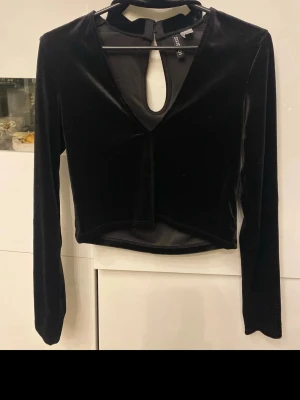 Svart sammets topp med nyckelhålsringning - Snygg svart topp i mjuk sammet med lång ärm och croppad passform. Toppen har en djup v-ringning och ett nyckelhålsformat hål framtill som ger en unik look. Perfekt för dig som vill sticka ut lite extra. Använd en gång 