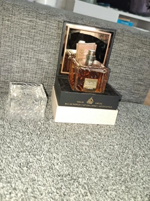 Khamrah Eau de Parfum från Lattafa - Khamrah Eau de Parfum från Lattafa i en elegant fyrkantig glasflaska med gulddetaljer. Flaskan har ett genomskinligt lock med mönster och kommer i en lyxig svart och beige förpackning. Doften är orientalisk och exklusiv, perfekt för dig som gillar unika parfymer.