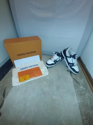 Louis Vuitton svarta och vita sneakers - Snygga Louis Vuitton sneakers i svart och vitt med klassiskt monogrammönster på sidorna. Skorna har vita snören, svarta detaljer och tydlig LV-logga. Tillverkade i en mix av läder och textil för en exklusiv känsla.