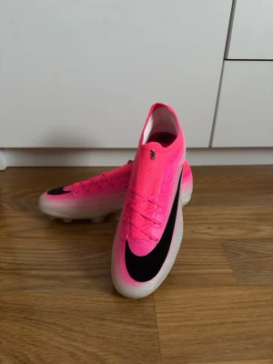 Nike Mercurial rosa fotbollsskor - Säljer ett par Nike Mercurial fotbollsskor i en riktigt snygg rosa färg med svart swoosh på ovansidan. Skorna har låg profil, snörning och är tillverkade i ett lätt syntetmaterial som ger bra bollkontroll och grepp på planen. Perfekta för dig som vill sticka ut på fotbollsplanen.