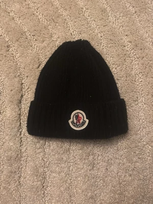  Moncler Mössa  - Säljer en svart ribbstickad mössa från Moncler med uppvikt kant och klassisk Moncler-logga framtill. Mössan är i mjukt material och har en enkel, stilren design som passar till det mesta.