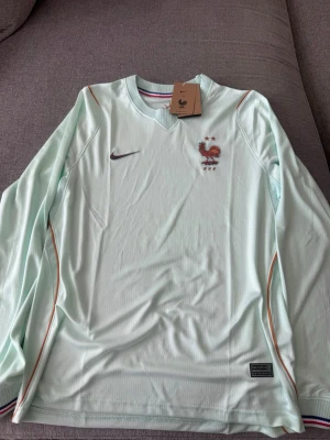 Ljusblå långärmad Nike FFF tröja - Snygg ljusblå långärmad tröja från Nike med FFF-logga och tupp på bröstet. Tröjan har diskreta ränder i orange och vitt längs axlarna och en ribbad krage med franska flaggans färger på insidan. Perfekt för fotboll eller träning.