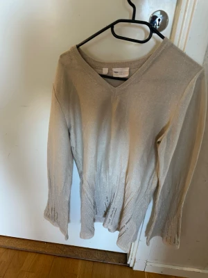 Beige stickad tröja med v-ringning - Beige stickad tröja från bpc med v-ringad hals och långa ärmar. Tröjan har ribbstickade muddar vid ärmslut och nederkant samt en lätt glansig finish i materialet. Perfekt för dig som gillar en enkel men ändå stilren look.