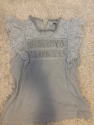 Ljusblå blus med volang från Lindex - Supersöt ljusblå blus från Holly & Whyte by Lindex med broderade volangärmar och rynkade detaljer framtill. Blusen har rund halsringning och knäppning bak i nacken. Perfekt för en romantisk och trendig look.