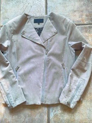 Beige skinnjacka med dragkedjor - Snygg beige mockajacka i bikerstil med asymmetrisk dragkedja framtill och dekorativa dragkedjor vid ärmslut och fickor. Jackan har en stilren design utan krage och tryckknappar på slaget. Perfekt för dig som vill ha en trendig och enkel look.