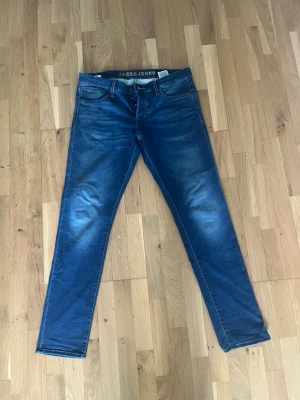Blå slim fit jeans från Jack & Jones - Snygga blå slim fit jeans från Jack & Jones med klassisk femficksmodell och lätt slitna detaljer på låren. Jeansen har normal midja och raka ben, perfekta till vardagslooken. Tillverkade i mjukt denimtyg för skön känsla.