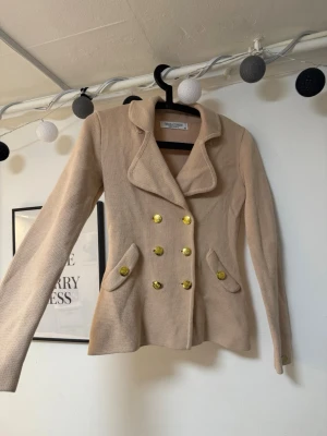 Beige kavaj med guldknappar - Stilren beige kavaj med markerad midja och breda slag. Dubbelknäppt framtill med sex blanka guldknappar och två snedställda fickor. Tillverkad i ett stickat material som ger en mjuk och bekväm känsla. Perfekt för en trendig och uppklädd look.