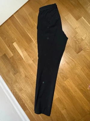 Svarta klassiska kostymbyxor - Säljer ett par svarta kostymbyxor med rak passform och enkel design. Byxorna har hög midja och är perfekta för en clean och stilren look. Materialet känns mjukt och följsamt, troligtvis polyester eller bomullsmix. Passar dig som vill ha ett par basic byxor till garderoben. Knappen för att stänga byxorna längst uppe är borta men om man har bälte så spelar det ingen roll och gör ingen skillnad 