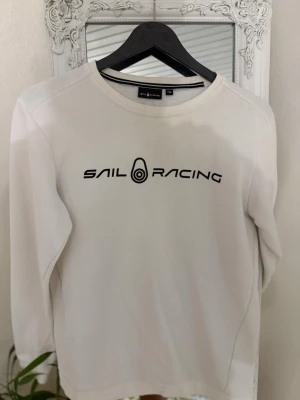 Vit sweatshirt från Sail Racing - Vit sweatshirt från Sail Racing med svart logotyp och text på bröstet. Tröjan har rund halsringning och långa ärmar. Tillverkad i mjukt material som passar perfekt till en avslappnad stil.