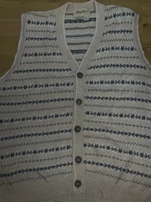 Beige stickad väst med mönster - Snygg beige stickad väst med grått och svart mönster, knäpps med fem knappar framtill. Djup V-ringning och ribbade kanter. Perfekt att slänga över en skjorta eller t-shirt för en trendig lager-på-lager-look.