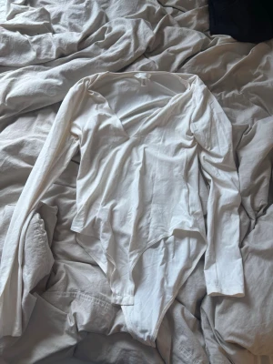 Vit långärmad body från H&M - Klassisk vit body från H&M med långa ärmar och djup v-ringning. Tillverkad i mjukt och stretchigt material som sitter skönt mot kroppen. Perfekt att styla med jeans eller kjol för en clean look.