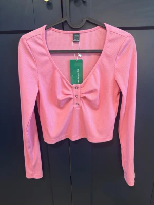 Helt ny Rosa ribbad topp från Shein - Trendig rosa ribbad topp från SHEIN med lång ärm och croppad passform. Toppen har en snygg v-ringning, dekorativa knappar framtill och en liten rosettliknande detalj vid bysten. Perfekt för dig som gillar en feminin och modern stil.