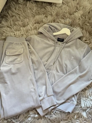 Grå velourset från Juicy Couture - Mjuk och lyxig grå velourset från Juicy Couture med dragkedja, huva och broderad logga på tröjan. Byxorna har ficka med knappdetalj. Perfekt för chill dagar hemma eller när du vill vara extra bekväm. Tröjan är i storlek L och byxorna i S