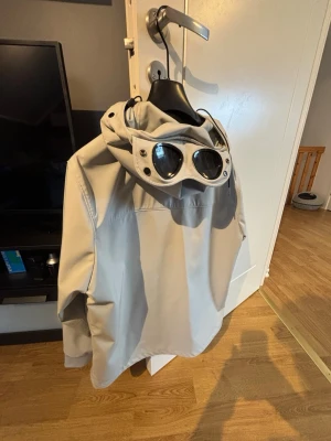Ljusgrå vindjacka med goggles-huva - (PRISET EJ HUGGET I STEN) nygg ljusgrå vindjacka med dragkedja framtill och två sidofickor med svarta dragkedjor. Jackan har en unik huva med inbyggda goggles och metallknappar, samt justerbara snören. Perfekt för dig som vill sticka ut med en modern streetwear-look.