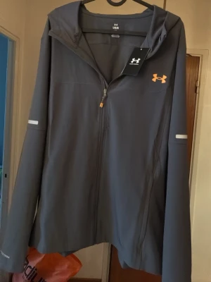 Grå vindjacka från Under Armour - Snygg grå vindjacka från Under Armour med huva och dragkedja framtill. Jackan har orange logga på bröstet och reflekterande detaljer på ärmarna. Tillverkad i ett lätt och smidigt material som passar perfekt för blåsiga dagar. skick 10/10