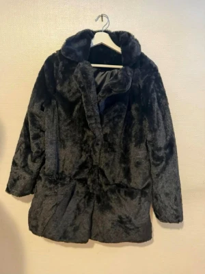 Svart fuskpälsjacka med krage - Säljer en svart fuskpälsjacka med bred krage och rak passform. Jackan har långa ärmar och stängs framtill. Perfekt för dig som vill ha en mjuk och varm look under vintern. Klassisk och enkel design som passar till många stilar.