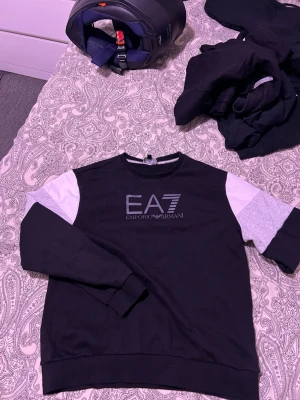 EA7 Emporio Armani svart set - Säljer ett svart set från EA7 Emporio Armani med matchande tröja och byxor. Setet har vita och grå detaljer på ärmar och ben samt logga på bröstet och benet. Mjukt material och avslappnad passform, perfekt för en chill look.