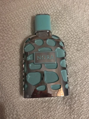 NORA parfymflaska i blått och silver - Unik parfymflaska med namnet NORA i mitten. Flaskan har en cool design med blå och silverfärgade detaljer i ett mönster som påminner om stenar. Locket är ljusblått och flaskan har en blank metallisk finish.