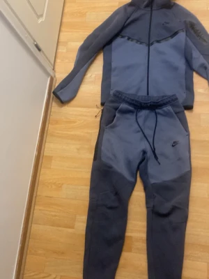 Nike Tech Fleece set mörkblå - Nike Tech Fleece set i mörkblå . Setet består av en zip-hoodie med hög krage och matchande byxor med dragsko i midjan. Båda plaggen har svarta detaljer och Nike-logga. Materialet är mjukt och stretchigt, perfekt för träning eller chill.