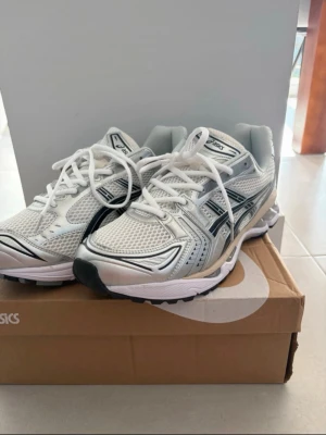 Asics gel kayano 14 skor  - Snygga vita och silverfärgade Asics sneakers med svart detalj på sidorna. Skorna har meshpaneler för bra ventilation och en chunky sula som ger skön dämpning. Klassisk sportig look med vita skosnören och logga på plösen. Perfekta för en trendig och bekväm stil.