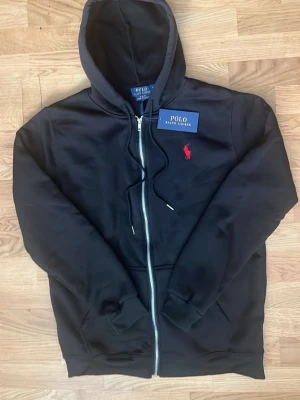 Svart hoodie från Polo Ralph Lauren - Svart hoodie med dragkedja från Polo Ralph Lauren. Klassisk design med huva och snören, samt röd broderad logotyp på bröstet. Mjukt material och ribbade muddar vid ärmslut och nederkant. Perfekt för en avslappnad och stilren look.