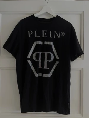 Svart Philipp Plein T-shirt - Logotyp, rundad hals, glitter, 100% bomull. Köpt från farfetch 2022, har tyvärr ej kvar kvitto. Nypris låg på 4500kr. Grymt skick och sitter något större än TTS