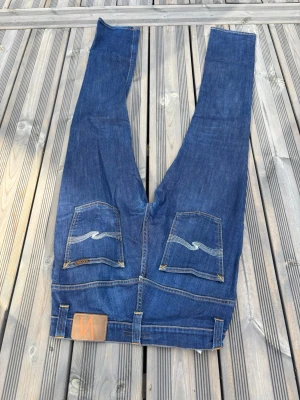 Blåa nudie jeans - Säljer ett par klassiska blå jeans med raka ben och snygga kontrastsömmar på bakfickorna. Jeansen har fem fickor och en läderpatch i midjan. Perfekta till vardags och passar till det mesta. Materialet är slitstark denim i bomull.