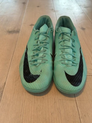 Turkosa futsalskor med svart swoosh - Snygga turkosa futsalskor med klassisk svart swoosh på sidorna. Skorna har snörning och låg profil, perfekt för snabba rörelser på planen. Tillverkade i syntetmaterial för lätt känsla och bra bollkontroll.