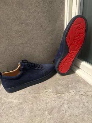 Mörkblå sneakers med röd sula - Snygga mörkblå sneakers i mocka från Christian Louboutin med den ikoniska röda sulan och svarta detaljer. Skorna har lågt skaft, matchande mörkblå snörning och beige insida. Perfekta för dig som vill sticka ut med exklusiv stil.