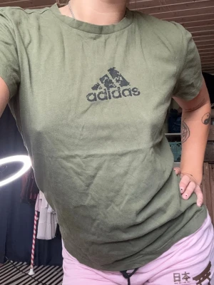 Olivgrön Adidas t-shirt - Snygg olivgrön t-shirt från Adidas med klassisk logga i svart framtill. T-shirten har rund halsringning och korta ärmar. Perfekt till jeans eller shorts för en avslappnad stil.Denna budas ut på min live 25 augusti 19.00