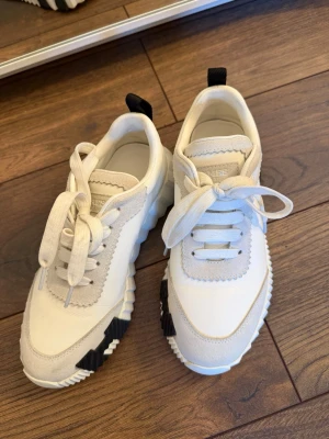 Vita sneakers med svarta detaljer H - Säljer ett par snygga vita sneakers med chunky sula och svarta detaljer på sidan och hälen. Skorna har snörning och en cool mix av material med både släta och mockaliknande partier. Perfekta för dig som gillar en modern och sportig look.