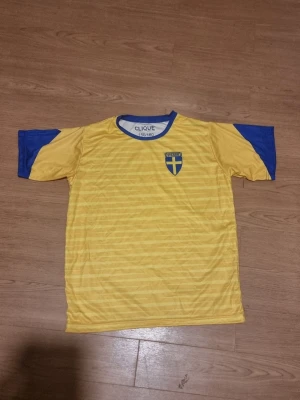 Sverige tröja - INSTABOX!!! Gul sportig t-shirt från Clique med blå detaljer på ärmar och krage. Bröstet har ett Sverige-emblem med blått och gult kors. Perfekt för träning eller att heja på landslaget. Tillverkad i lätt och snabbtorkande material.