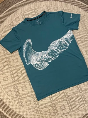 Nike bone running tshirt - Nike Dri-Fit t-shirt i blått med korta ärmar och en stor vit grafisk skelett print på framsidan. Perfekt för löpning eller träning, tillverkad i ett lätt och snabbtorkande material. Klassisk Nike-logga på ärmen och rund halsringning. Passform S/M Junior 160-172 cm ish