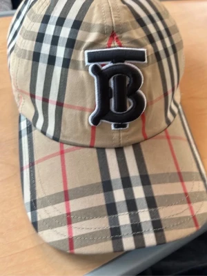Burberry keps - Säljer en beige keps från Burberry med det klassiska rutiga mönstret i svart, vitt och rött. Kepsen har en böjd skärm och ett broderat TB-monogram framtill. Tillverkad i ett lätt och bekvämt material som passar perfekt till streetwear och trendiga outfits.