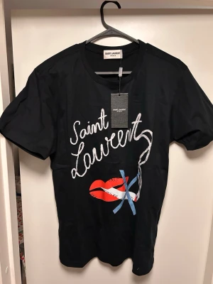 Svart Saint Laurent t-shirt med tryck - Svart t-shirt från Saint Laurent med coolt tryck framtill i vitt, rött och blått. T-shirten har rund hals och korta ärmar. Tillverkad i mjuk bomull för en skön känsla och snygg passform.