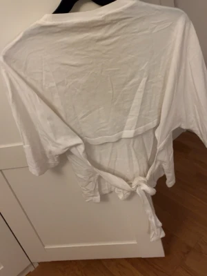 Vit omlottopp med knytband och öppen rygg  - Vit topp i mjukt material med korta ärmar och knytband i midjan och öppen rygg. Omlottmodell som ger en avslappnad och trendig look. Perfekt att styla med jeans eller kjol för en enkel men snygg outfit. Passar Xs-L beroende på hur man knyter